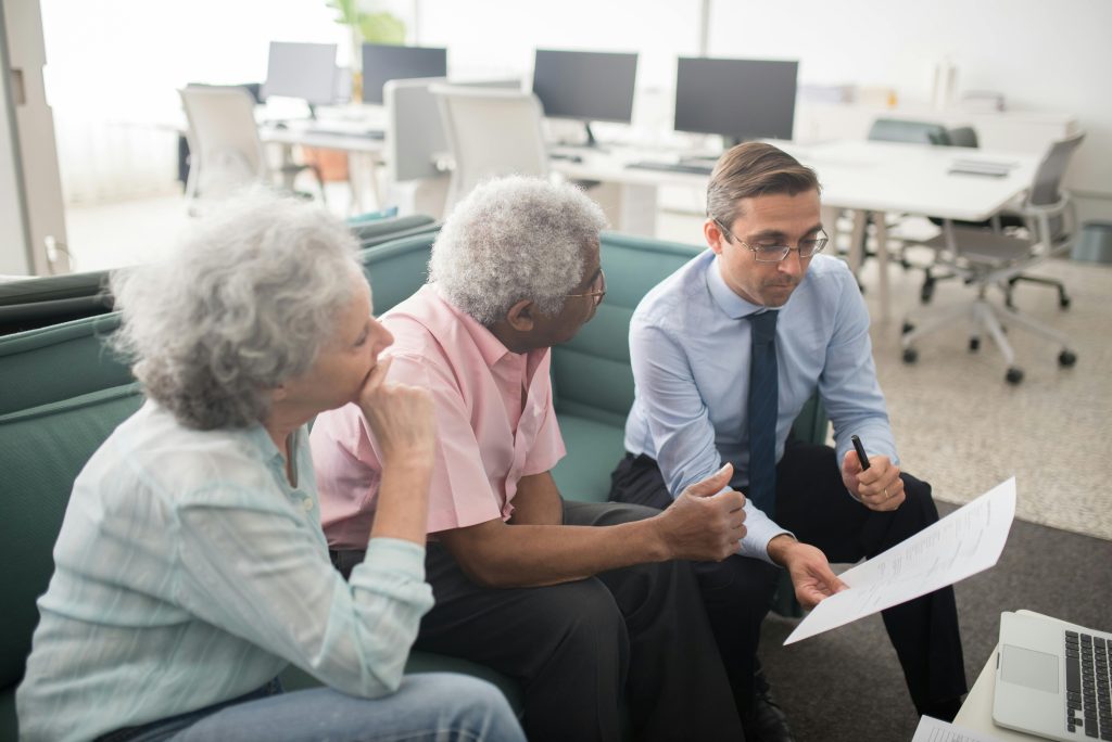 Business professional consults elderly clients in an office setting. Collaborative discussion, paperwork visible.